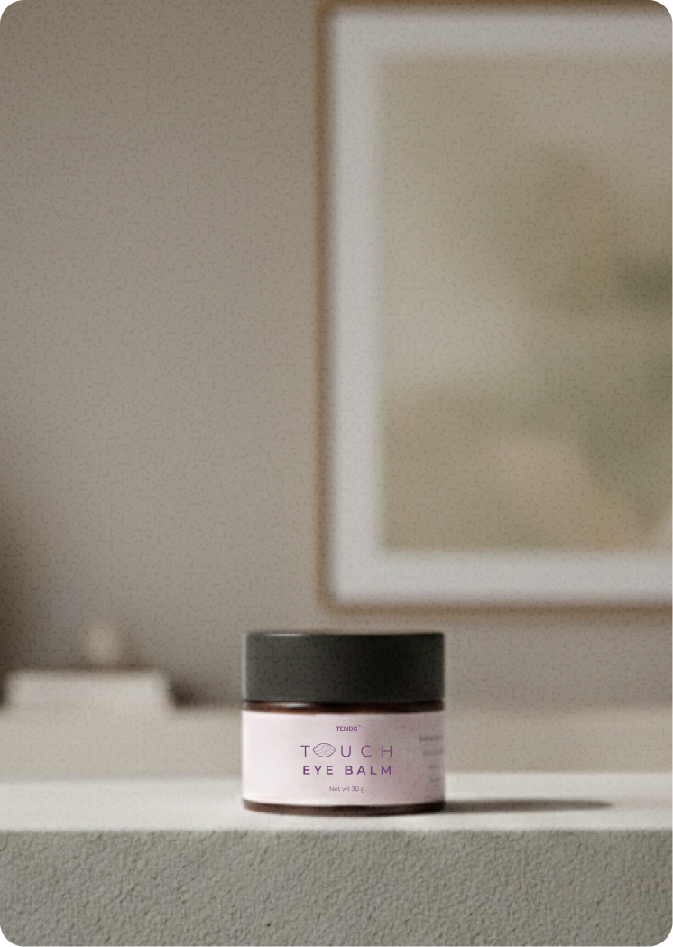 TENDS Touch Eye Balm jar with a black lid placed on a neutral surface in a softly lit indoor setting, with a blurred framed artwork in the background.