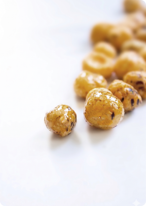 One golden-coated air-popped lotus seed snack in the foreground, a loose cluster of the same snacks behind it, on a plain white surface — small, round, and whole. 