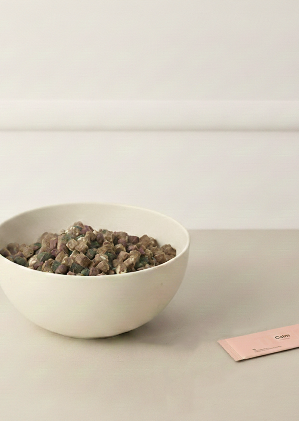 Beige ceramic bowl filled with dry dog food on a neutral surface, with a pink sachet labeled “Calm” placed nearby.
