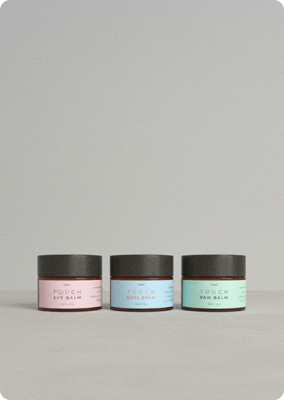 Three TENDS Touch balm jars with black lids arranged side by side on a neutral surface, labeled Eye Balm, Nose Balm, and Paw Balm in pink, blue, and green.