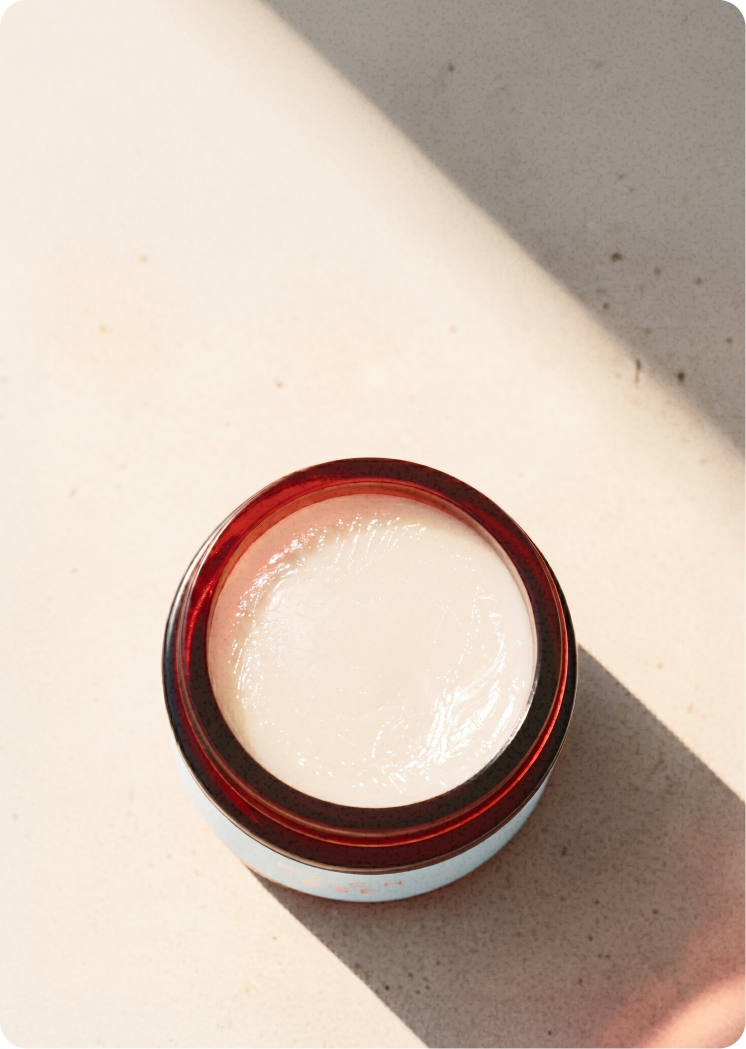 Top-down view of an open amber glass jar containing a smooth, cream-coloured balm with a soft swirl on the surface, lit by strong natural light casting a shadow.